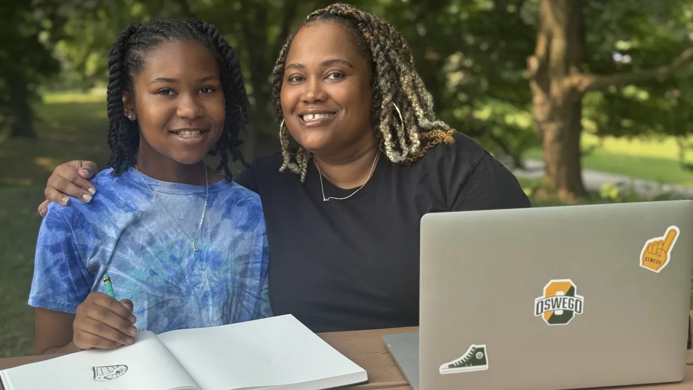 female adult learner in the park pose with her daughter while she works on her laptop